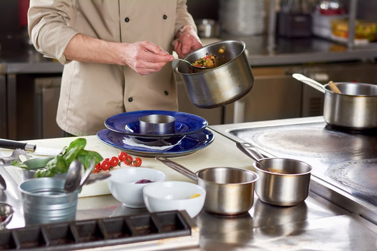 Chef put out with spoon stewing vegetables. Cropped image of male chef going putting garnishing into stainless bowl. Food preparation at professional kitchen.