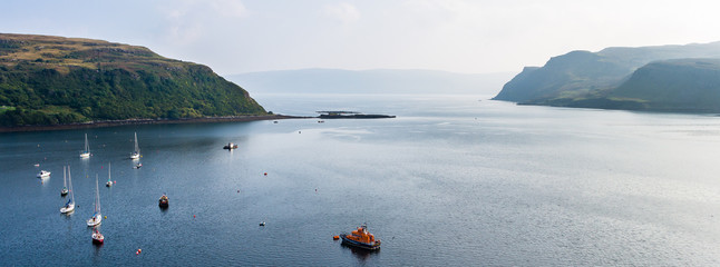 Panorama of moored fishermen boats at Loch Portree waters in Portree's harbor, in the Isle of Skye (Scotland, UK) with the dimmed silhouette of Raasay in the background in a sunny day.