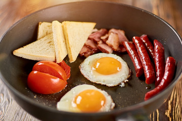 Hot english breakfast in skillet. Fried eggs with yolks, baked sausages and ham, toasts and tomatoes. Fresh english breakfast.