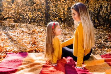 Happy smiling mother and daughter in yellow pullovers sitting on the wool plaid in the autumn park. Happy family outdoors. Autumn park. Leaf fall.