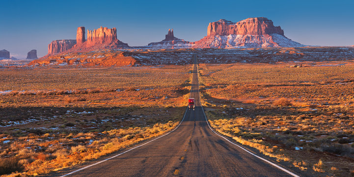 Red car driving on a straight road in Monument Valley early on a Sunny morning.