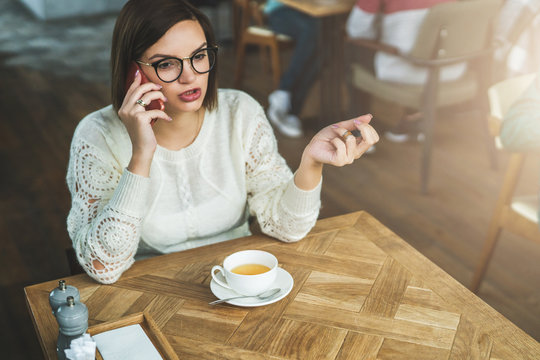 Young businesswoman in glasses and white sweater is sitting in cafe at wooden table and talking on mobile phone. Telephone conversations. Hipster girl relaxing, drinking tea, calling her friends.