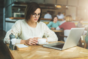 Young businesswoman sitting in cafe at table,drinking coffee and working on laptop.Student is studying online.Hipster girl using computer.Online education, marketing, e-learning.Social media, network.