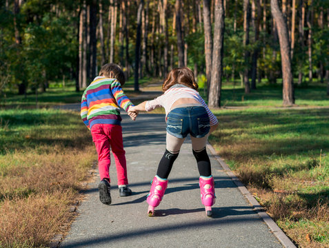 Boy helps the girl to roller-skate in the park. Brother supports his sister, who is riding on the rollers.