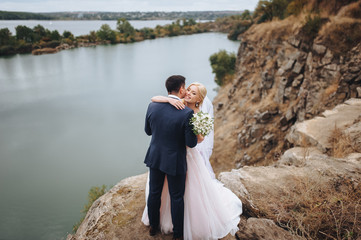 The bride and groom stand on the river bank against the rocks and gently embrace. Wedding concept.