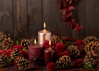 Three candles of crimson and pink color on a dark background with cones, leaves and daisies.