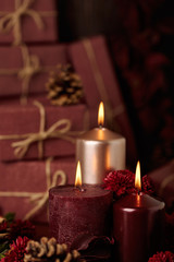 Three candles of crimson and pink color on the background of gift boxes with cones, leaves and daisies.