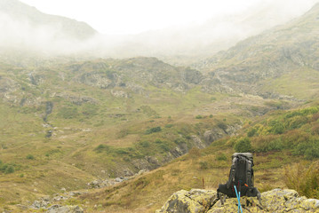 A view of a mountain valley covered with clouds. In the foreground on the stone is a backpack, to the stone are propped poles for the nordic walk