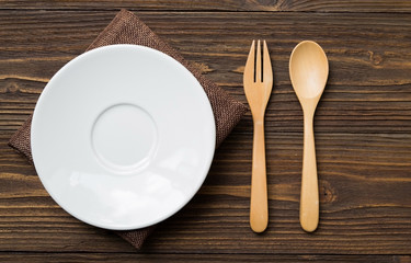 A white plate rests on a brown cloth. There are spoon and fork made of wood placed next to it. All were put on an old brown wood table.
