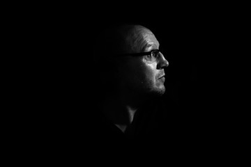 Side profile of a white middle aged man with spectacles or glass in shadow against a black background looking up towards the light