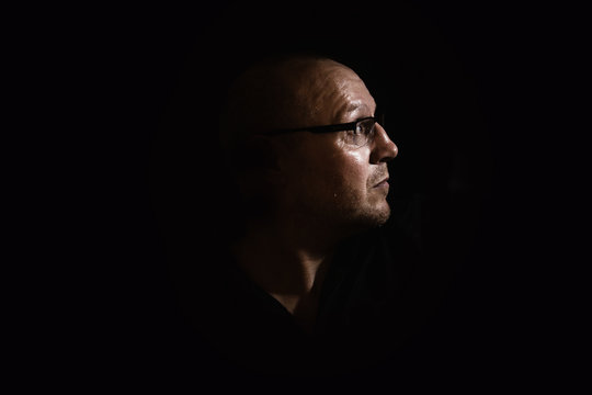 Side profile of a white middle aged man with spectacles or glass in shadow against a black background looking up towards the light