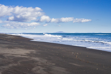 Fototapeta na wymiar Little waves of Pacific ocean are broken on the shore. Splashes of it goes on a black volcano sand surface of a beach. Kamchatka. Russian Federation. October of 2017. Light blue sky.