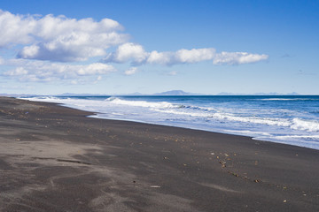 Little waves of Pacific ocean are broken on the shore. Splashes of it goes on a black volcano sand surface of a beach. Kamchatka. Russian Federation. October of 2017. Light blue sky.