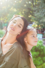 Young mother and daughter sitting back to back in nature in sunlight