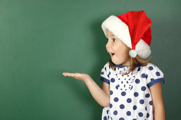 surprised child girl with santa hat holds his hand near empty school blackboard, copy space