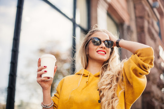 The concept of street fashion. young stylish girl student wearing boyfrend jeans, white sneakers bright yellow sweetshot.She holds coffee to go. portrait of smiling girl in sunglasses