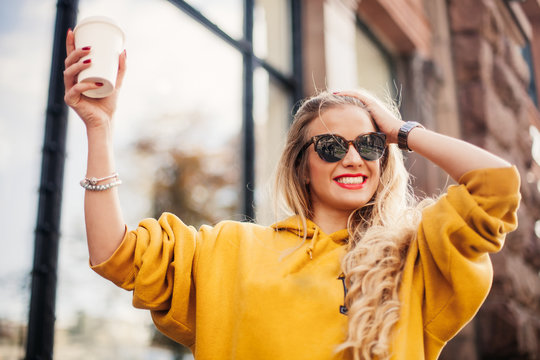 Stylish happy young woman wearing boyfrend jeans, white sneakers bright yellow sweetshot.She holds coffee to go. portrait of smiling girl in sunglasses
