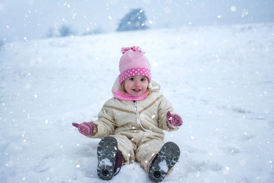 Happy little girl playing with snow while sitting in winter outfit outdoors.