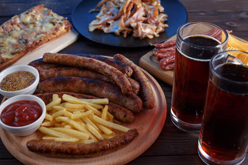 Oktoberfest food, appetizing beer snacks set for big company. Close up of two mugs of stout and grilled sausages with french fries on wooden table