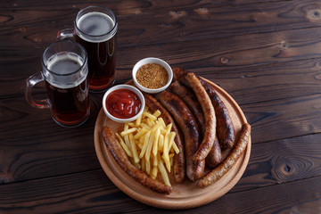 Dark beer and appetizing beer snacks set. Top view on table with two mugs of stout, wooden board with grilled sausages and french fries served with sauces. Oktoberfest food, pub concept