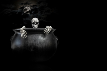 Halloween skeleton peaking out from the inside of a witches cauldron under a nice moon covered with dark clouds.