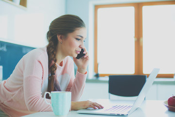 Young woman sits at the kitchen table using a laptop and talking on a cell phone.