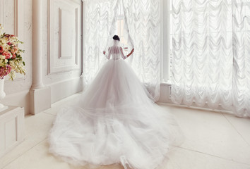 The bride stands at the window in a dress with a long train and veil.