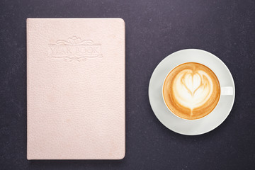 Hot latte coffee in white cup and notebook with blank empty cover on black stone table background