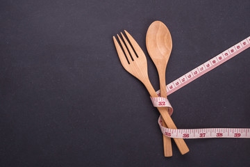 Measurement tape with wooden spoon and fork on black stone board. Eating control or diet concept