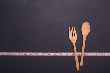 Measurement tape with wooden spoon and fork on black stone board. Eating control or diet concept