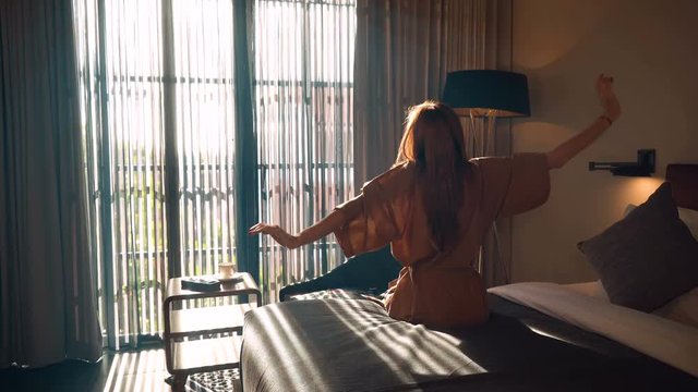 Young caucasian woman in brown beige robe stretches out hands sitting on neat bed in the morning in front of big window. Shot with Sony a7s and Atomos Ninja Flame.