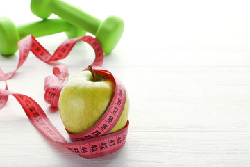 Measuring tape with green apple and dumbbells on white wooden table