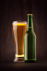 Green beer bottle and glass weizen with golden lager on dark brown wood board, vertical,mock up. Template for advertising, design, branding identity.