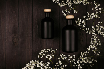 Blank black cosmetics bottles with white small flowers on dark wood board, mock up, top view....