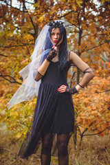 Fototapeta na wymiar Halloween composition. Beautiful dark haired girl in black dress witches costume with walking in the forest. Halloween Witch in a dark forest. Halloween art design. Background For Halloween Concept