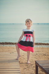 Young cute baby girl enjoying childhood summer time on sandy beach posing on wooden pier bridge wearing casual stylish dress
