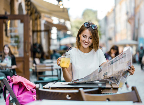 Pretty fair hair caucasian tourist girl with sunglasses on her head is having break with lemonad drink and city map in a street cafe, sunny day