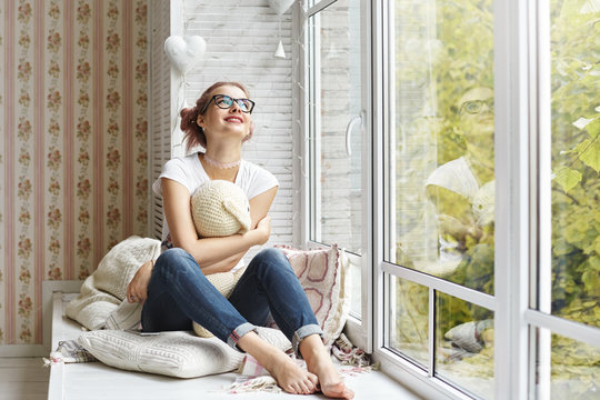 Teenage, Joy, Happiness And Leisure Time Concept. Childish 14 Year Old Teenage Girl Wearing Glasses, Choker And No Socks Embracing Her Plush Bunny While Daydreaming On Windowsill In Her Room