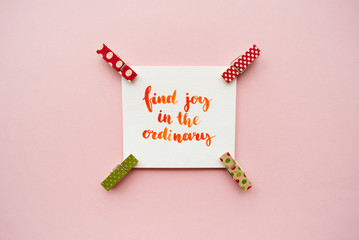Inspirational quote "find joy in the ordinary" handwritten with watercolor in calligraphy style, miniature clothespins on a pale pink pastel background
