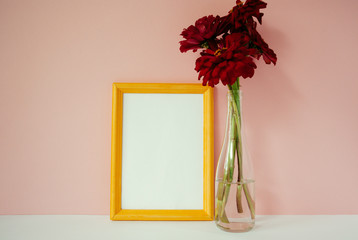 Yellow wooden photo frame mockup, red cynicism in glass vase in front of pale pink pastel background