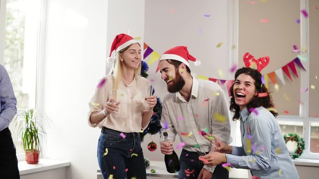Group of young people celebrating christmas and new year in the office opening a bottle of sparkling wine and squib petards with confetti. Slowmotion shot