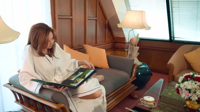Young woman in bathrobe on couch looking through photo album takes coffee