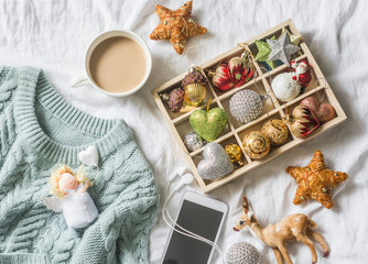 Christmas background. Box of vintage christmas decorations, coffee with milk, phone and blue knitted sweater on the bed, view from above. Christmas cozy mood still life