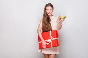 Happiness blonde woman in red cap holding champagne glass and gift box, looking at camera and toothy smiling.