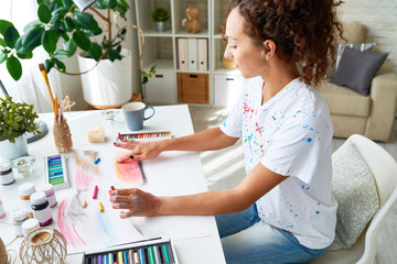 Portrait of young female artist finger painting with pastel colors sitting at modern desk at home