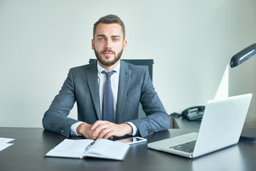 Confident bearded entrepreneur wearing classical suit looking at camera while sitting at desk, interior of modern office on background, waist-up portrait shot