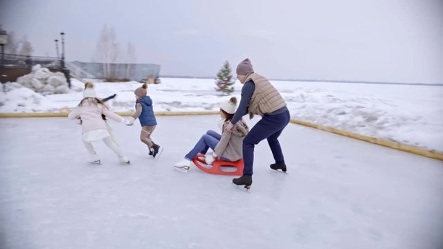 Tracking of happy little children and man skating on ice rink and pulling sled with cheerful young woman on nice winter day