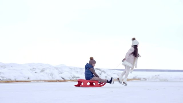 Tracking of adorable little girl wearing pink coat with warm hat and scarf skating on ice and pulling cute brother on sled, then spinning on rink