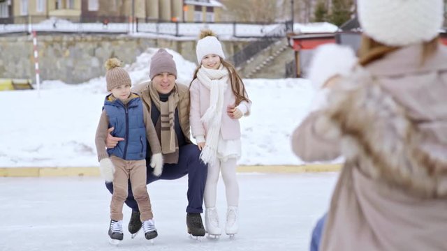 Over the shoulder shot of unrecognizable woman holding mobile phone and taking photo of cheerful man and little children in skates posing on outdoor ice rink in winter