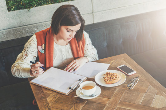Young businesswoman is sitting in cafe at table, signing documents. Student is studying online, doing homework. Girl is writing letter, statement. On table cup of tea, cookies, smartphone. E-learning.